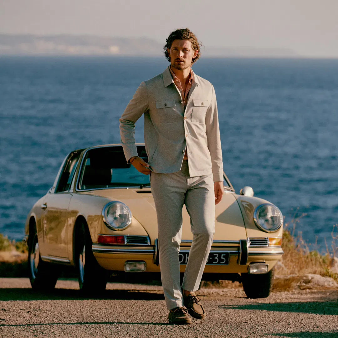 Man in beige overshirt en lichte pantalon bij een klassieke auto aan de kust